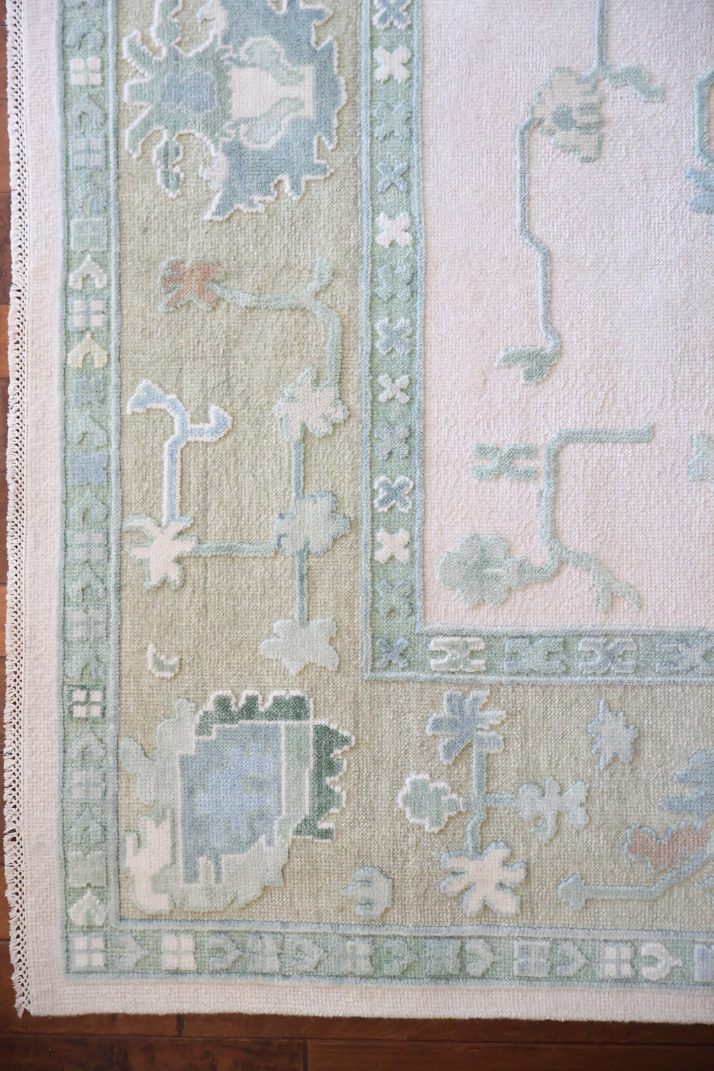 A close-up of a patterned area rug corner showing a cream field, pale sage green border, and muted blue and gray motifs.