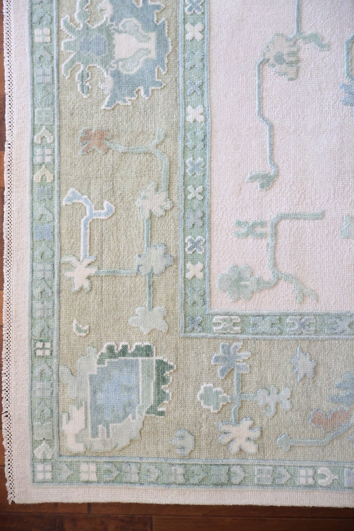 A close-up of a patterned area rug corner showing a cream field, pale sage green border, and muted blue and gray motifs.